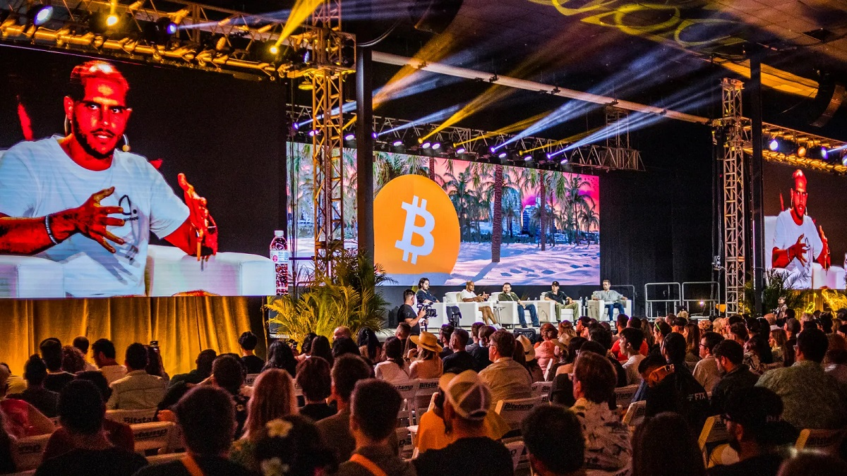 A large audience at a Bitcoin conference watching a panel of speakers on a stage with large screens displaying the Bitcoin logo.